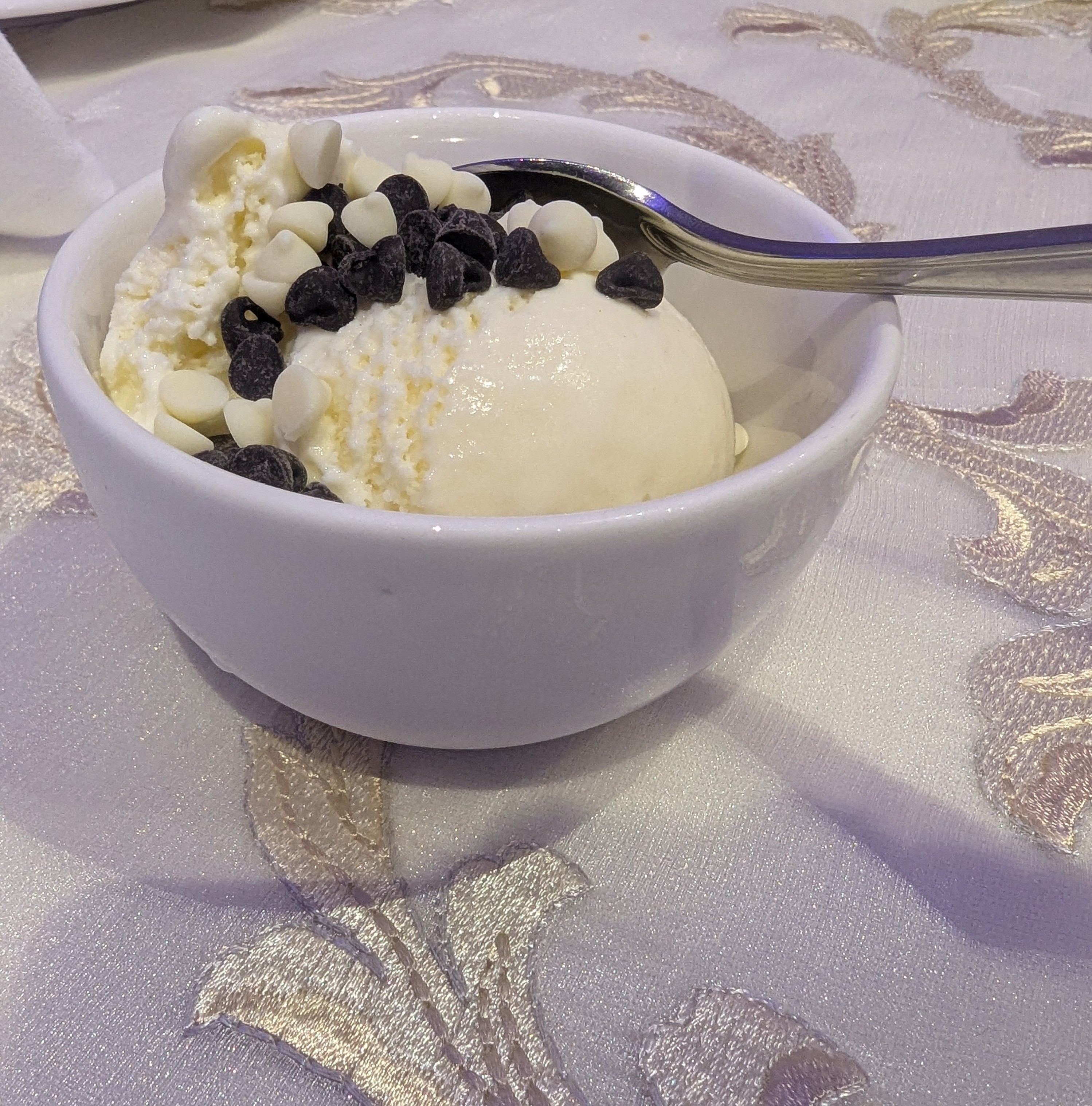 A bowl of vanilla ice cream topped with chocolate and white chocolate chips sits on a decorative tablecloth.