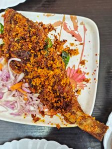 
A beautifully presented plate featuring a whole fried fish topped with a crispy, spiced coating. Surrounding the fish are slices of red onion and grated carrots, adding a pop of color