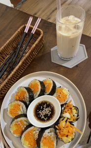 Top-down view of kimbap rolls arranged around a small bowl of soy sauce topped with sesame seeds, with iced milk tea and chopsticks on a dark wooden table.