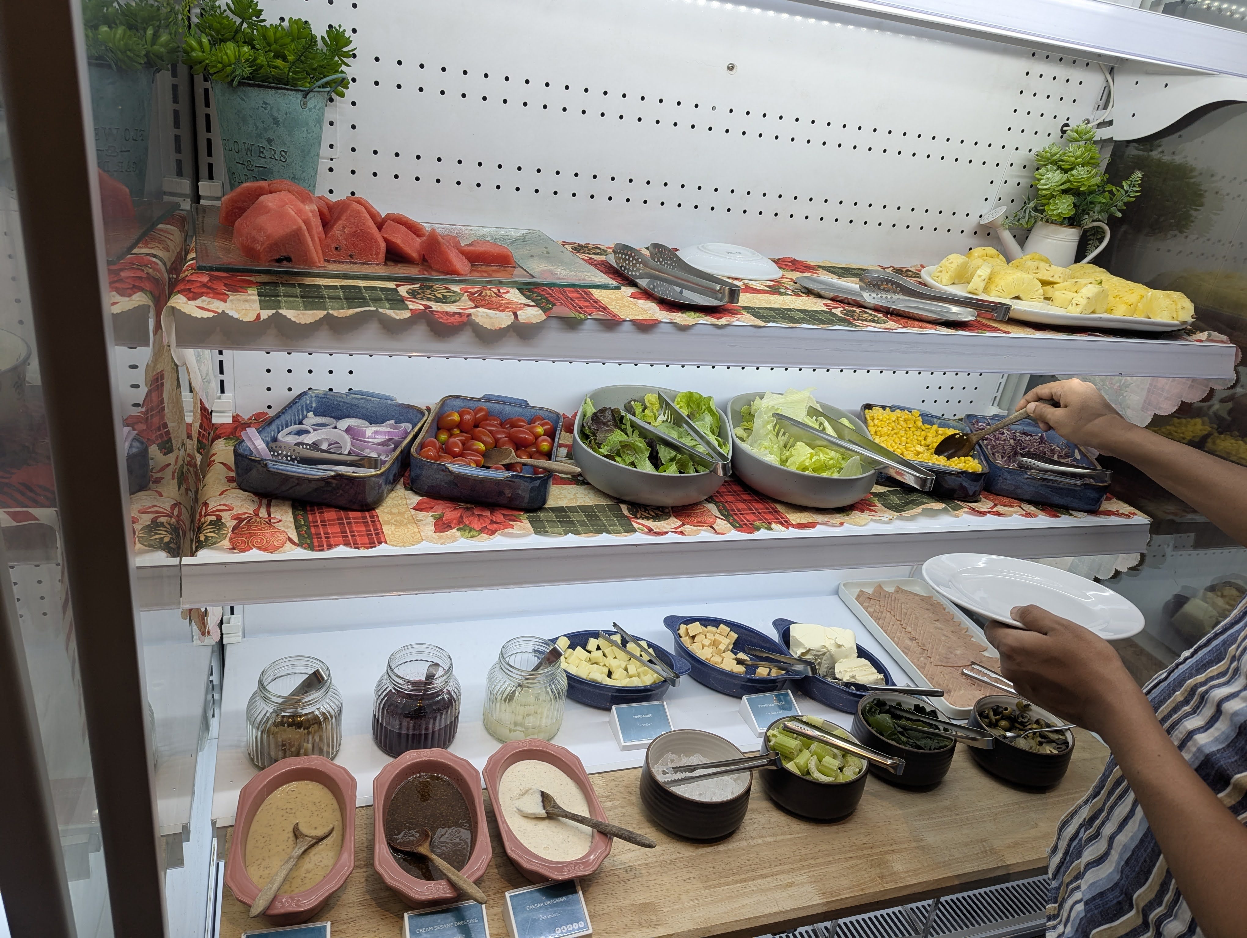 A well-stocked salad bar buffet with three shelves displaying sliced watermelon, pineapple, cherry tomatoes, mixed greens, corn, cheese cubes, and assorted dressings in jars, with a person serving themselves on the right