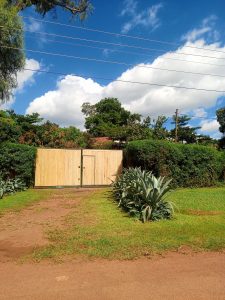 A wooden gate surrounded by trimmed hedges, agave plants, and grass under a blue sky with clouds.