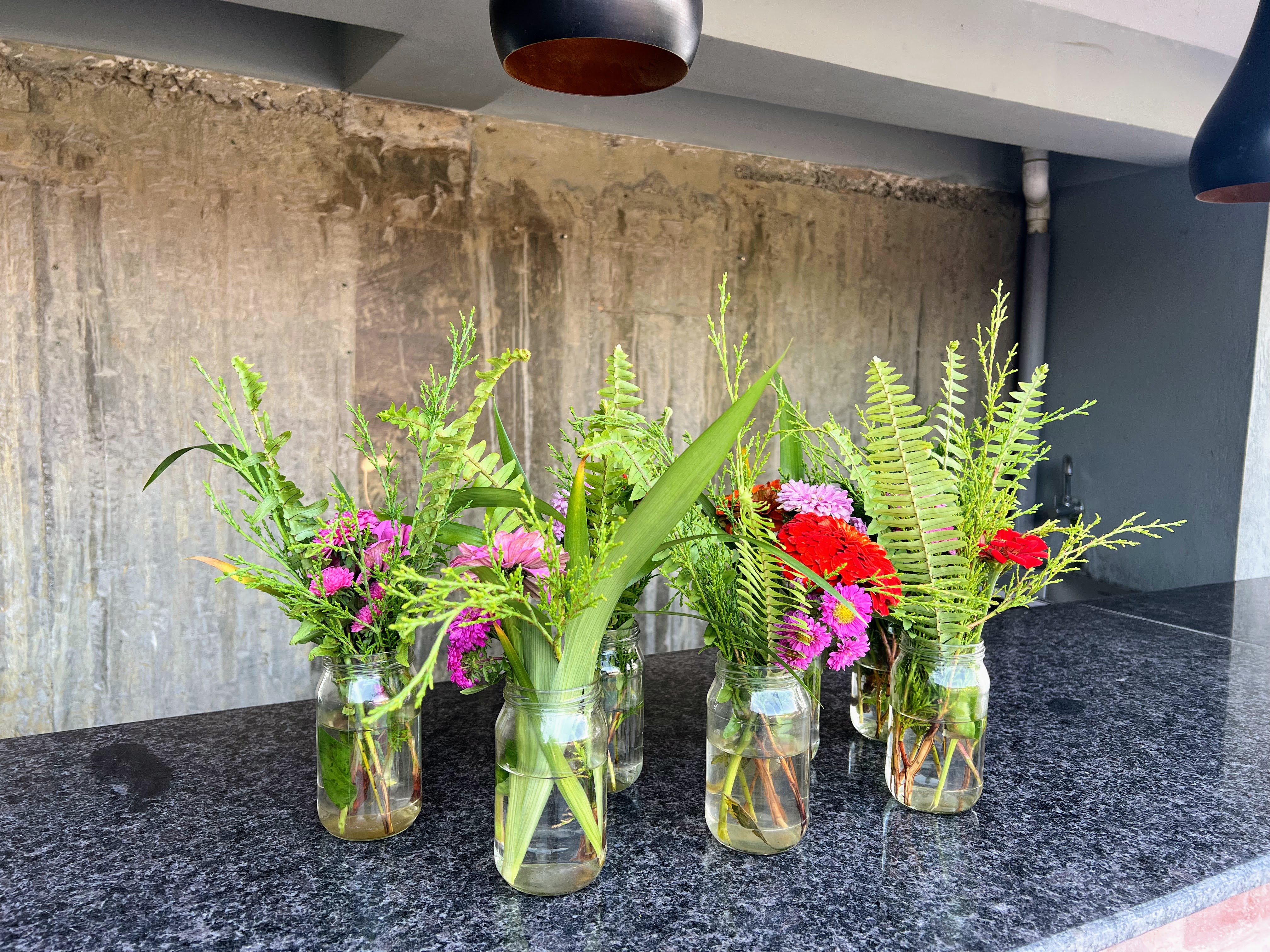 A row of small glass jars used as vases, filled with colorful flowers and green ferns, arranged on a dark countertop.