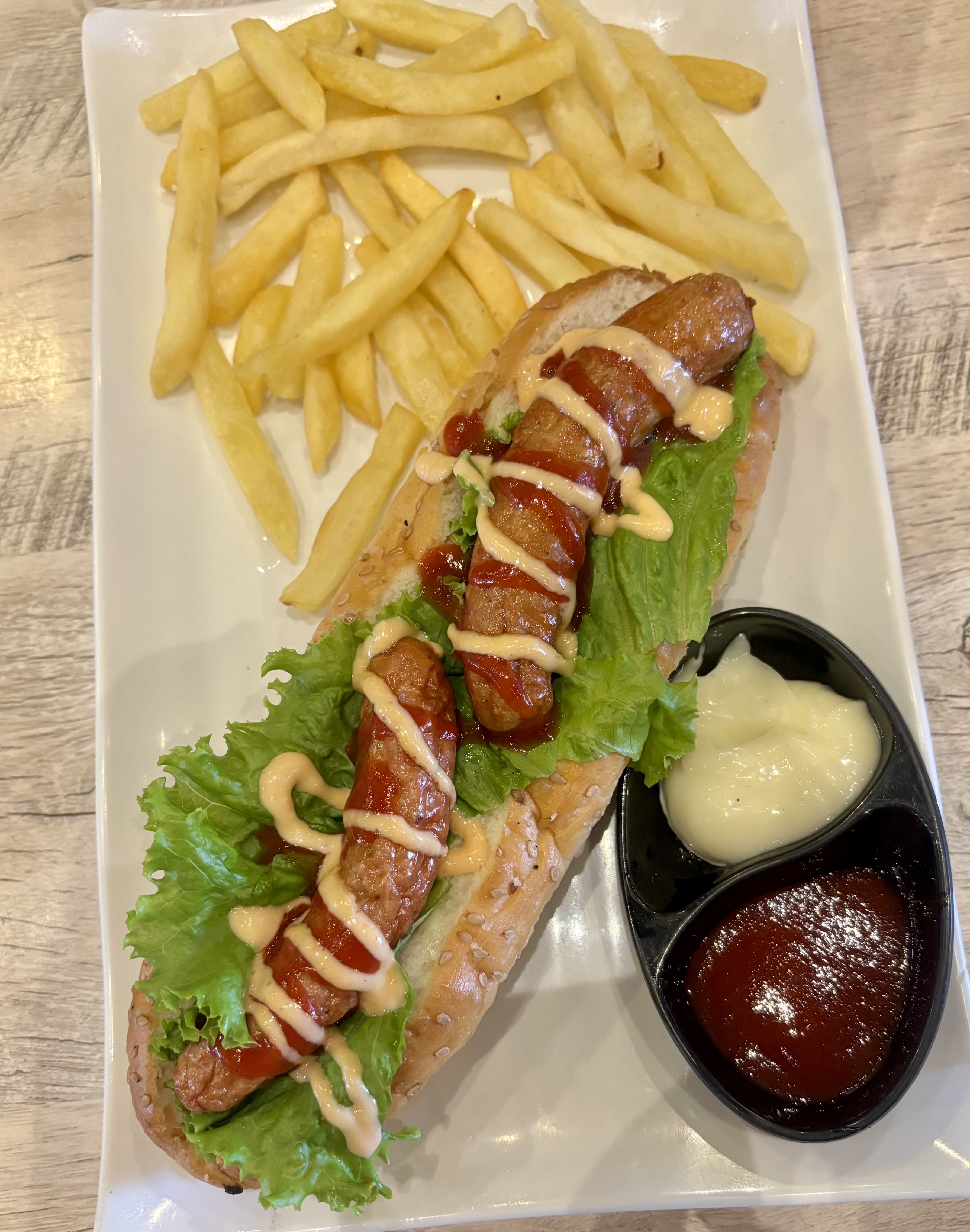 A hot dog with sausages, drizzled with mustard and ketchup, on lettuce in a bun, is served with fries and sauces on a white plate.