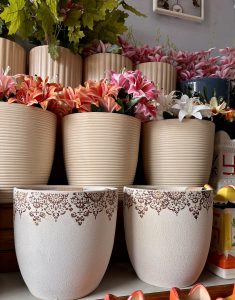

Decorative beige pots with intricate patterns are filled with vibrant pink, orange, and white flowers cascading down a shelf against a soft gray wall.