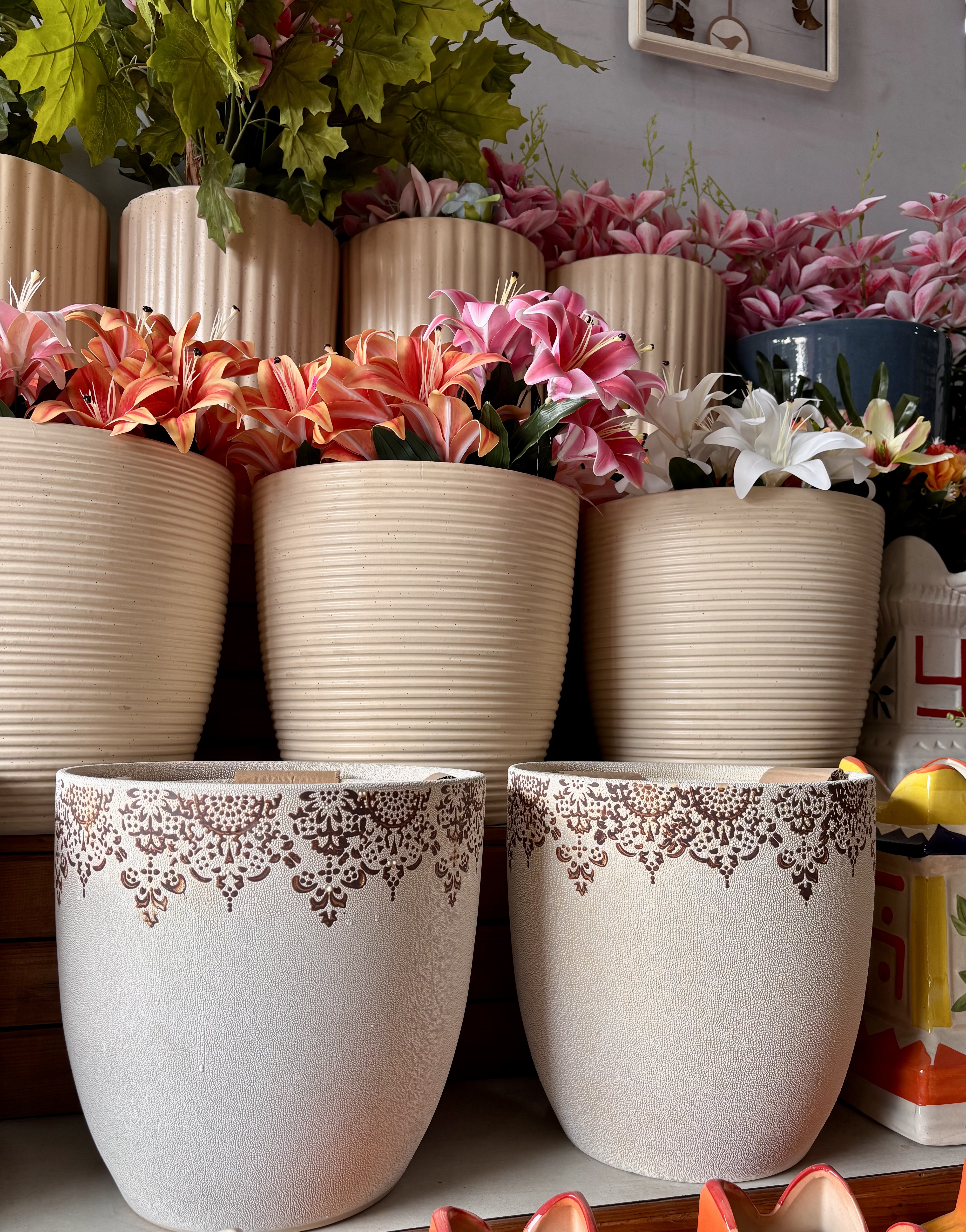 Decorative beige pots with intricate patterns are filled with vibrant pink, orange, and white flowers cascading down a shelf against a soft gray wall.
