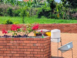 A red brick planter filled with vibrant red flowers and green leaves sits in a sunny garden surrounded by lush greenery.