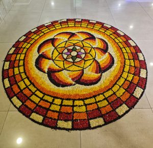
A vibrant circular rangoli design made from colorful rice and flower petals is displayed on a polished floor. 