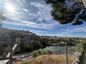 Scenic landscape with a winding river, a stone bridge, and a historic fortified structure under a blue sky.
