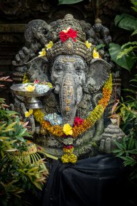 A statue of the Hindu god Ganesh, decorated with colorful flowers, surrounded by lush green plants.
