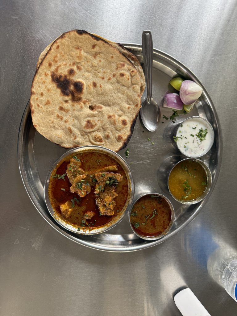 A metal plate holds a Varhadi Chicken Thali with tandoori roti, spicy chicken curry, yellow lentils, and a cooling yogurt sauce.