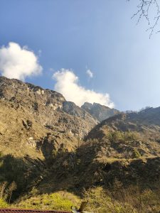 Rocky mountain landscape under a clear blue sky with a few fluffy white clouds, the sun casting a warm glow over rugged terrain and sparse greenery.