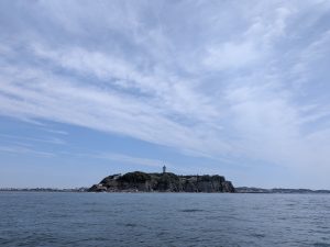 A rocky, green-covered island with a lighthouse stands in calm blue waters under a sky with wispy clouds. The view is of Enoshima Island seen from Sagami Bay, Japan.
