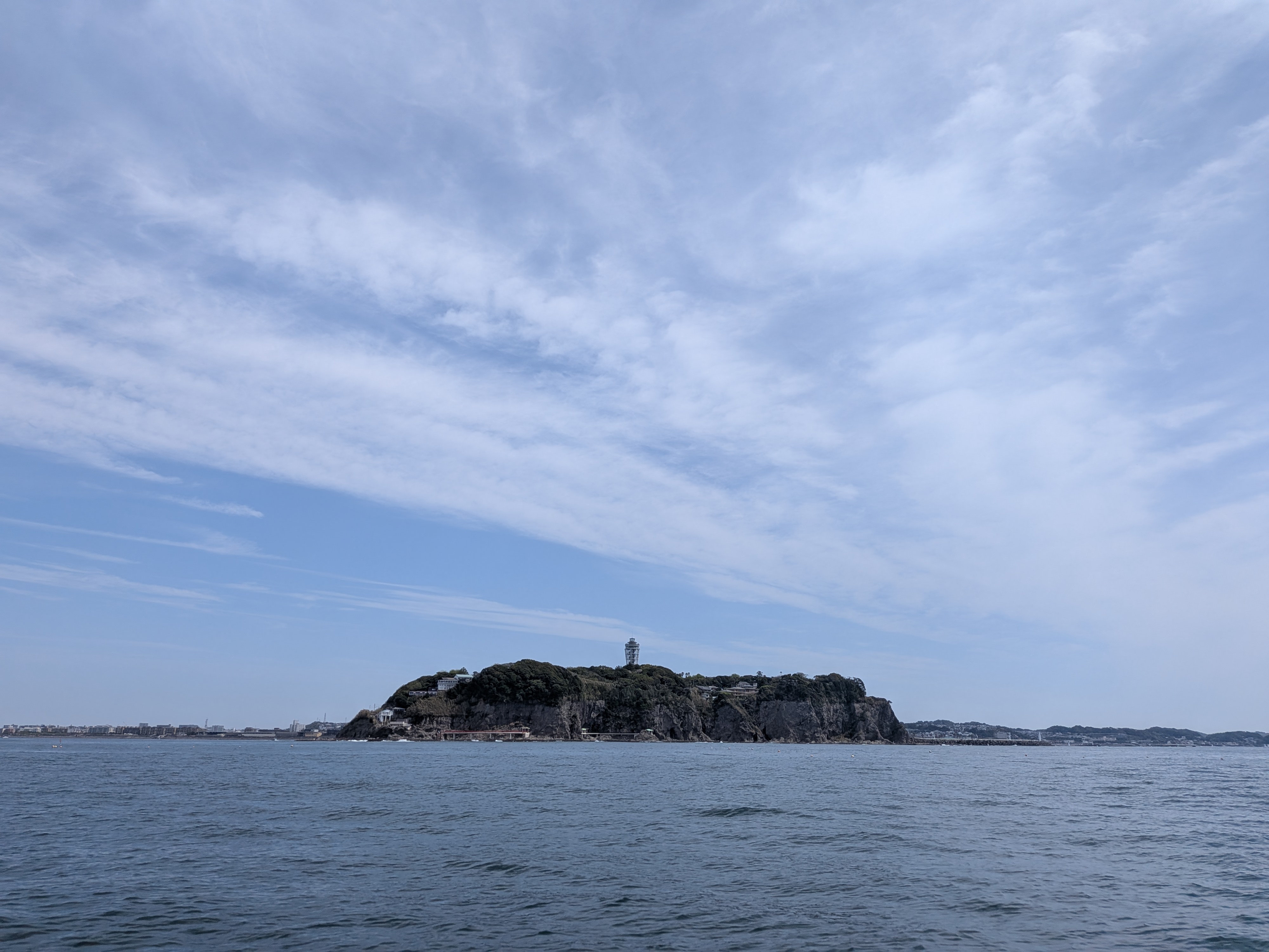 A rocky, green-covered island with a lighthouse stands in calm blue waters under a sky with wispy clouds. The view is of Enoshima Island seen from Sagami Bay, Japan.