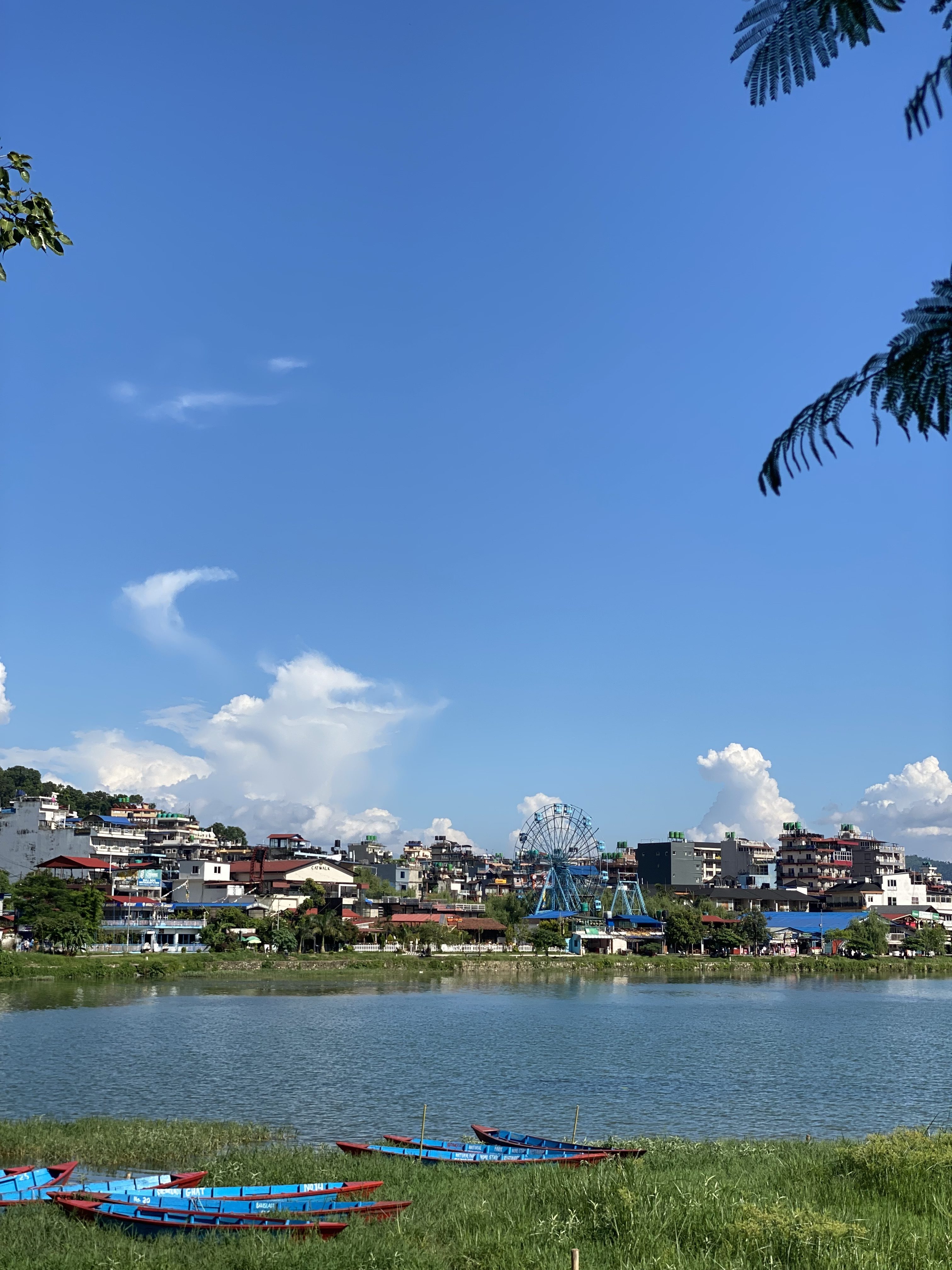 Calm Fewa lake with small blue boats on the shore, a town at the base of green hills, and a blue Ferris wheel under a bright sky with scattered clouds.