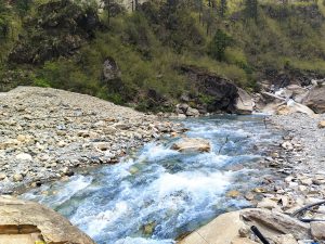 A clear blue river flowing swiftly over rocks in a mountainous landscape, surrounded by greenery and rocky slopes.