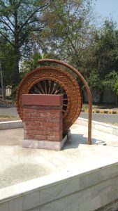 A decorative water-wheel sculpture, with intricate carvings, is mounted on a brick pedestal. The wheel is partially covered with a curved metal pipe. The surrounding area is landscaped with trees and greenery, and a road is visible in the background. 