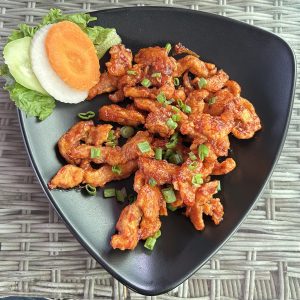 Spicy, orange-hued chicken strips garnished with chopped green onions on a black triangular plate, with sliced carrot, cucumber, and lettuce on the side.