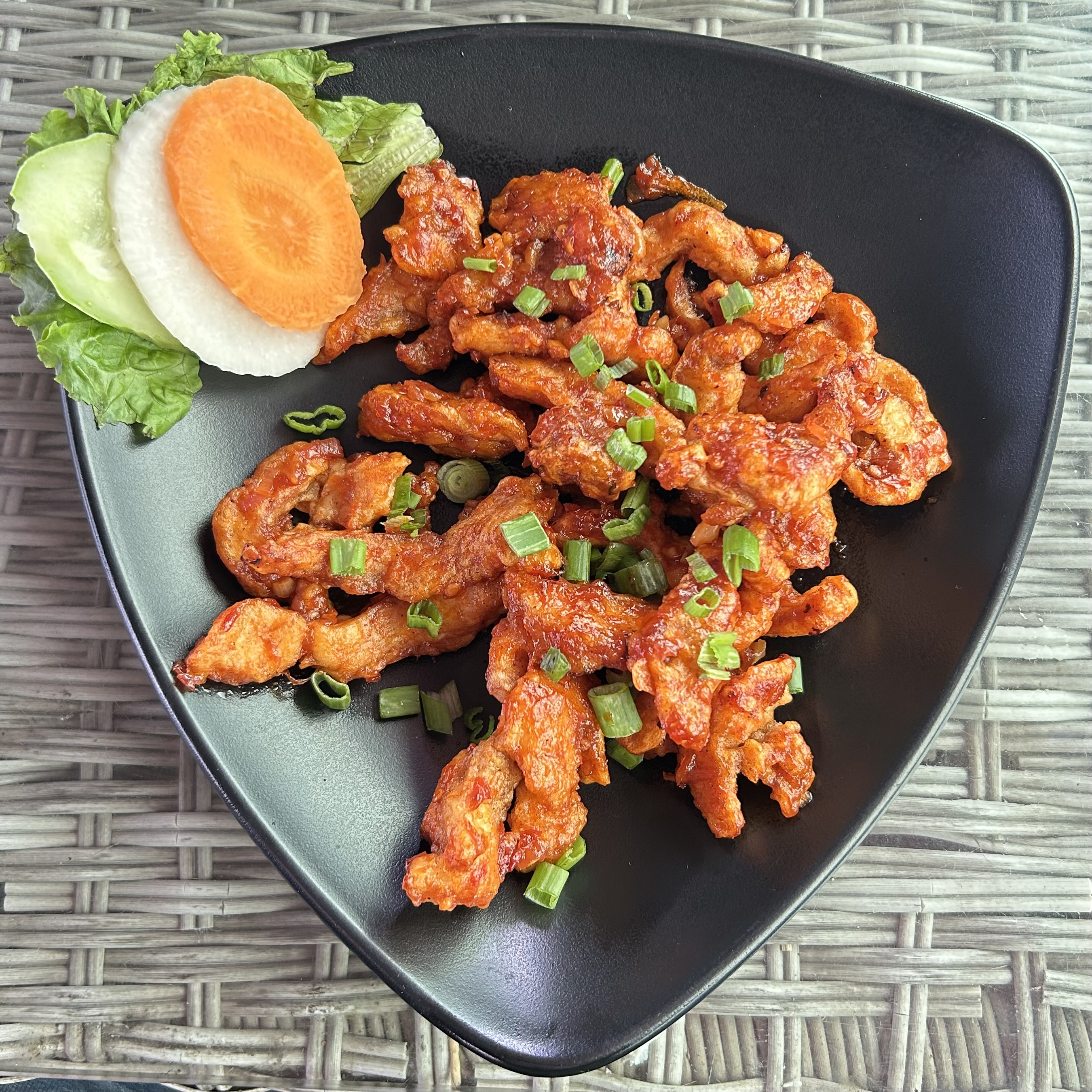Spicy, orange-hued chicken strips garnished with chopped green onions on a black triangular plate, with sliced carrot, cucumber, and lettuce on the side.