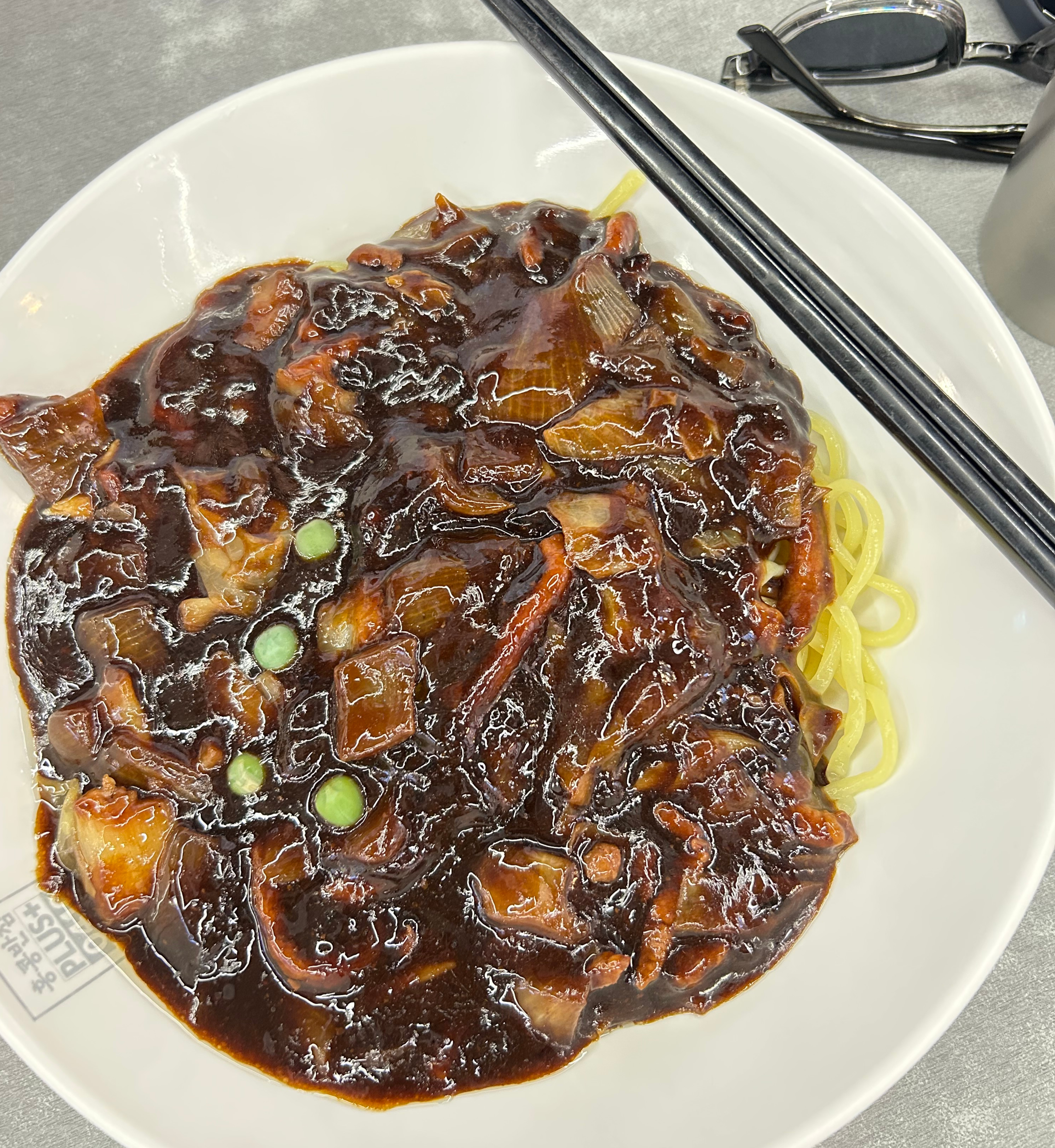 A white plate topped with noodles covered in rich, dark-brown sauce, garnished with green peas.