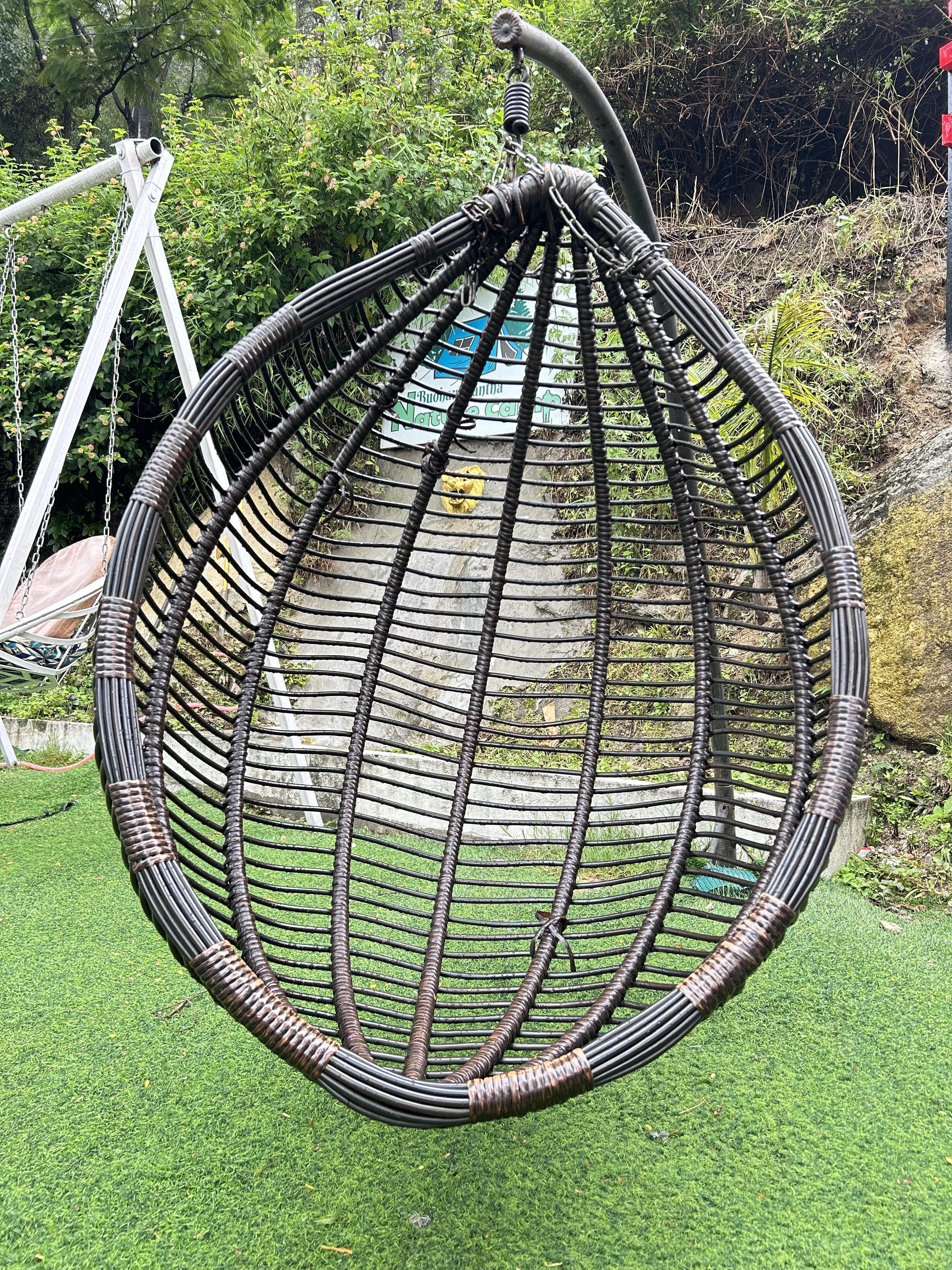 Hanging a wicker chair with brown tones on a green artificial turf. Surrounded by lush greenery and a playground swing in the background.