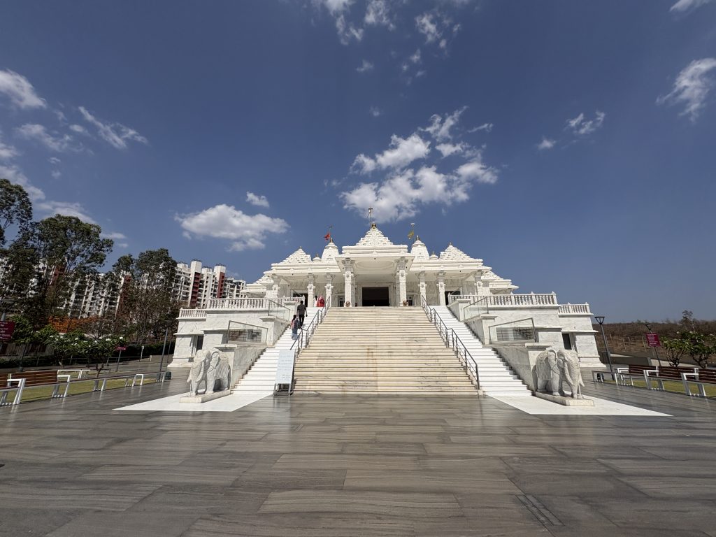 The image features wide angle view of a grand Trimandir Pune temple structure made of white stone, situated on a raised platform. It has ornate architectural details, including intricately carved pillars and tiered roofs. A flight of wide stairs leads up to the temple entrance, which is flanked by two large elephant sculptures.