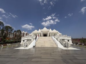 The image features wide angle view of a grand Trimandir Pune temple structure made of white stone, situated on a raised platform. It has ornate architectural details, including intricately carved pillars and tiered roofs. A flight of wide stairs leads up to the temple entrance, which is flanked by two large elephant sculptures.
