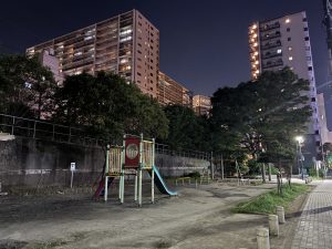 A quiet nighttime playground sits beside illuminated apartment buildings, bordered by trees and a sidewalk lined with bollards.