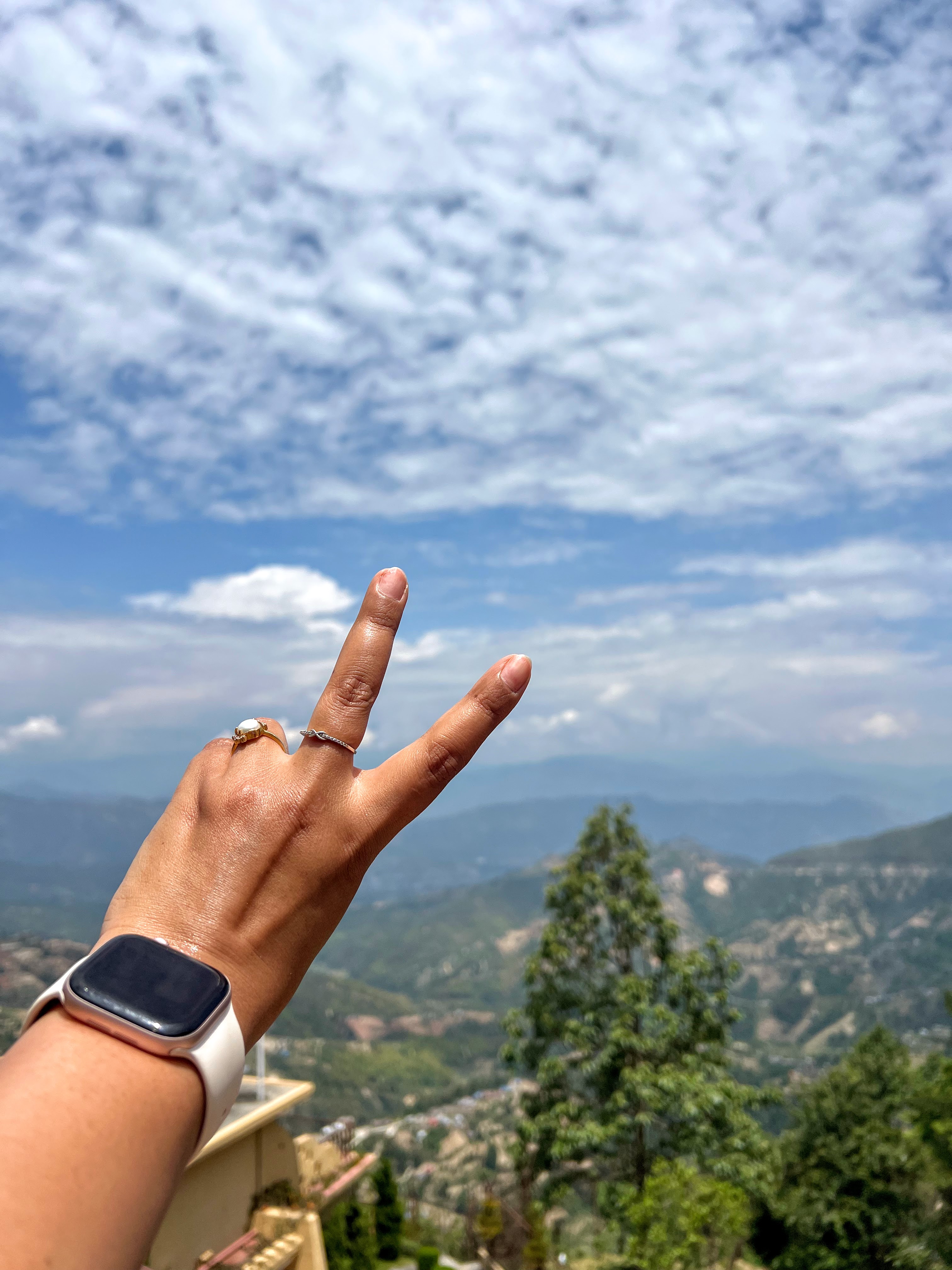 A first-person perspective of a hand making a peace sign against a background of green rolling hills and a blue sky with fluffy white clouds.