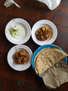 A wooden table displaying three white plates alongside a blue basket. One plate contains a serving of yogurt with sliced cucumbers and onions, another plate holds a vegetable curry with potatoes and peas, and the third plate features pieces of chicken in a rich sauce. The blue basket is filled with several flatbreads.