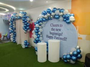 A balloon arch of silver, blue, and white surrounds a farewell backdrop reading “Cheers to the new beginnings! Happy Farewell!” in a bright and cheerful setting.