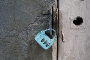 A small, turquoise-colored combination padlock hangs from a rusty metal latch on a weathered door. The padlock shows signs of wear, with some of its paint chipped away and a combination of three dials .