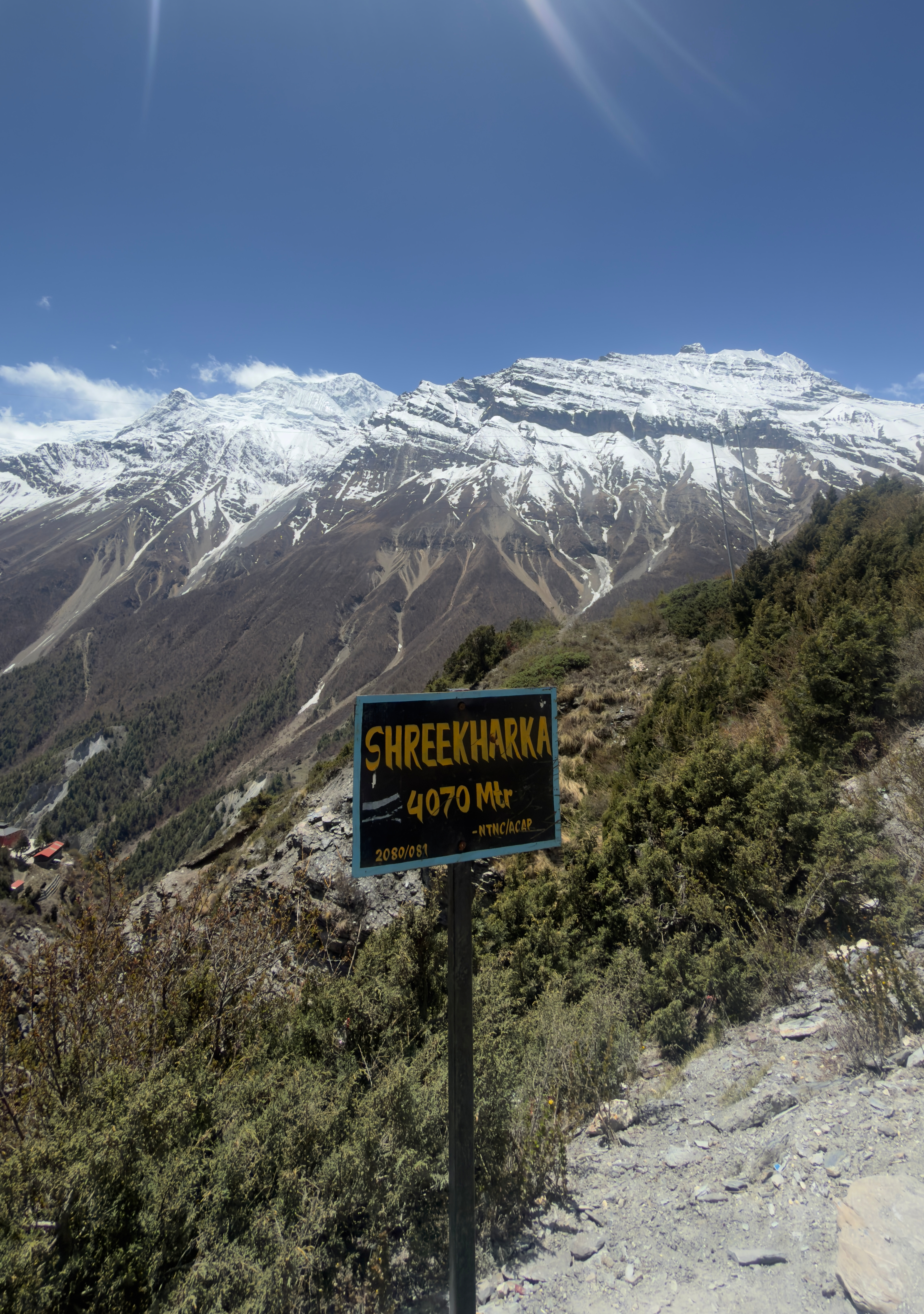 A sign reading "Shree Kharka 4070 Mtr" stands against a stunning backdrop of snow-capped mountains and clear blue sky, conveying an adventurous vibe.