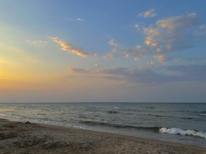 A serene beach scene at sunset, showcasing a gently lapped shore with fine sand in the foreground. The sun is low on the horizon, casting warm hues of orange and pink across the sky, blending with soft blues and hints of golden clouds. In the distance, a few small silhouettes of people can be seen enjoying the water, creating a peaceful and calming atmosphere.