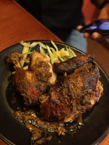 Grilled chicken pieces on a black plate with a glossy, spicy marinade, served with sliced cucumbers