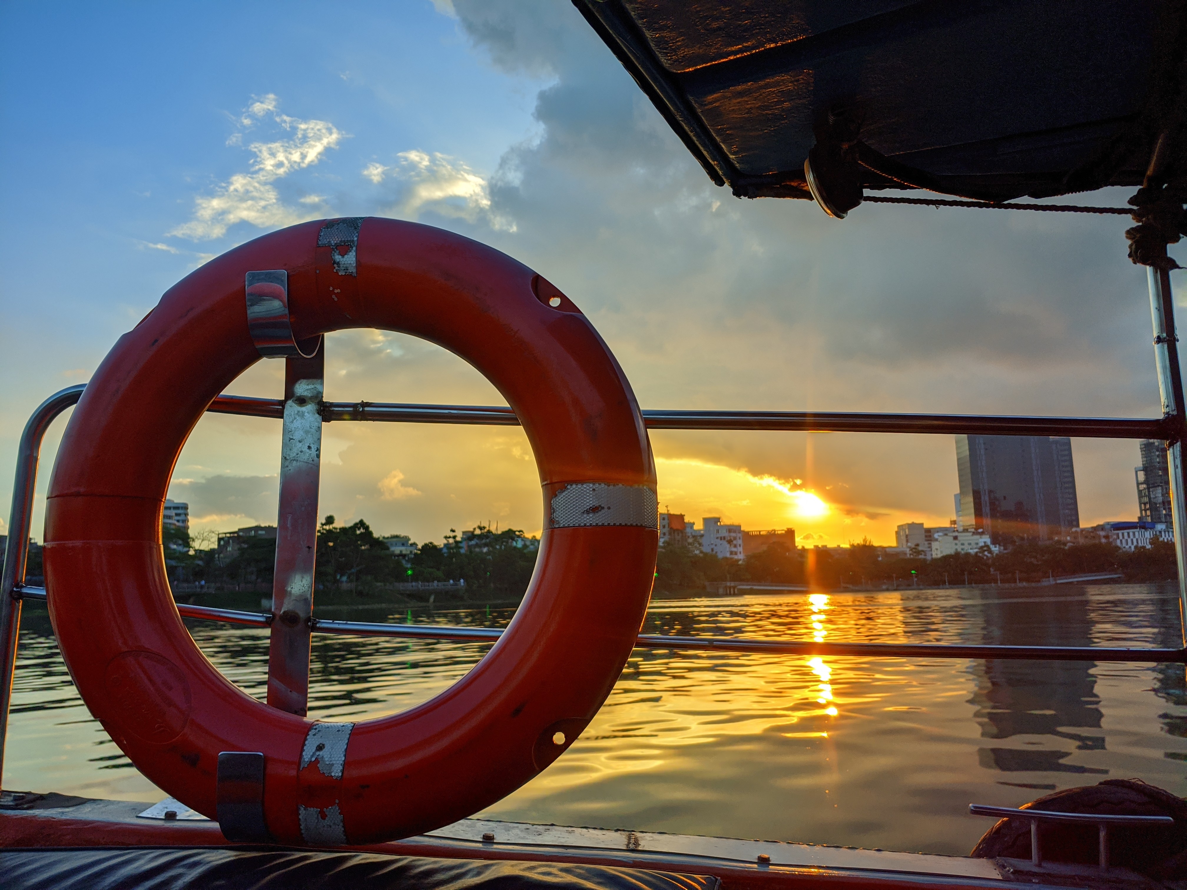 Sunset over calm water viewed from a boat with a red lifebuoy in the foreground. The sky is a mix of blue and orange hues, reflecting tranquility.