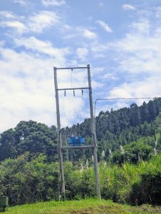 An electric transformer mounted on wooden poles in a lush green rural landscape, with trees in the background and a partly cloudy blue sky, creating a calm countryside scene.