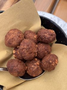 A serving of golden brown fried meatballs piled in a kraft paper lined black pan with a spoon underneath.
