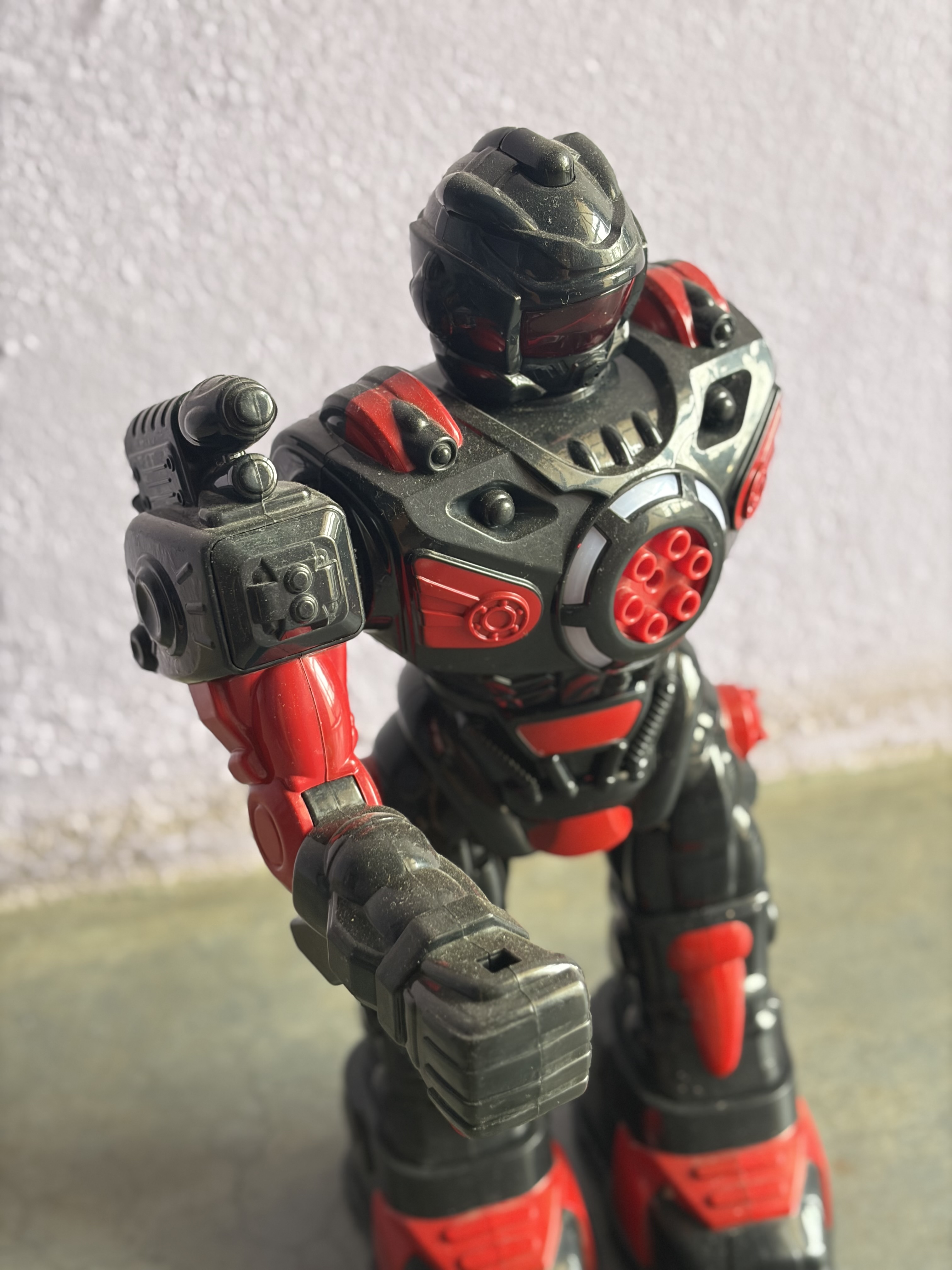 A toy robot with black and red armor stands against a textured wall.