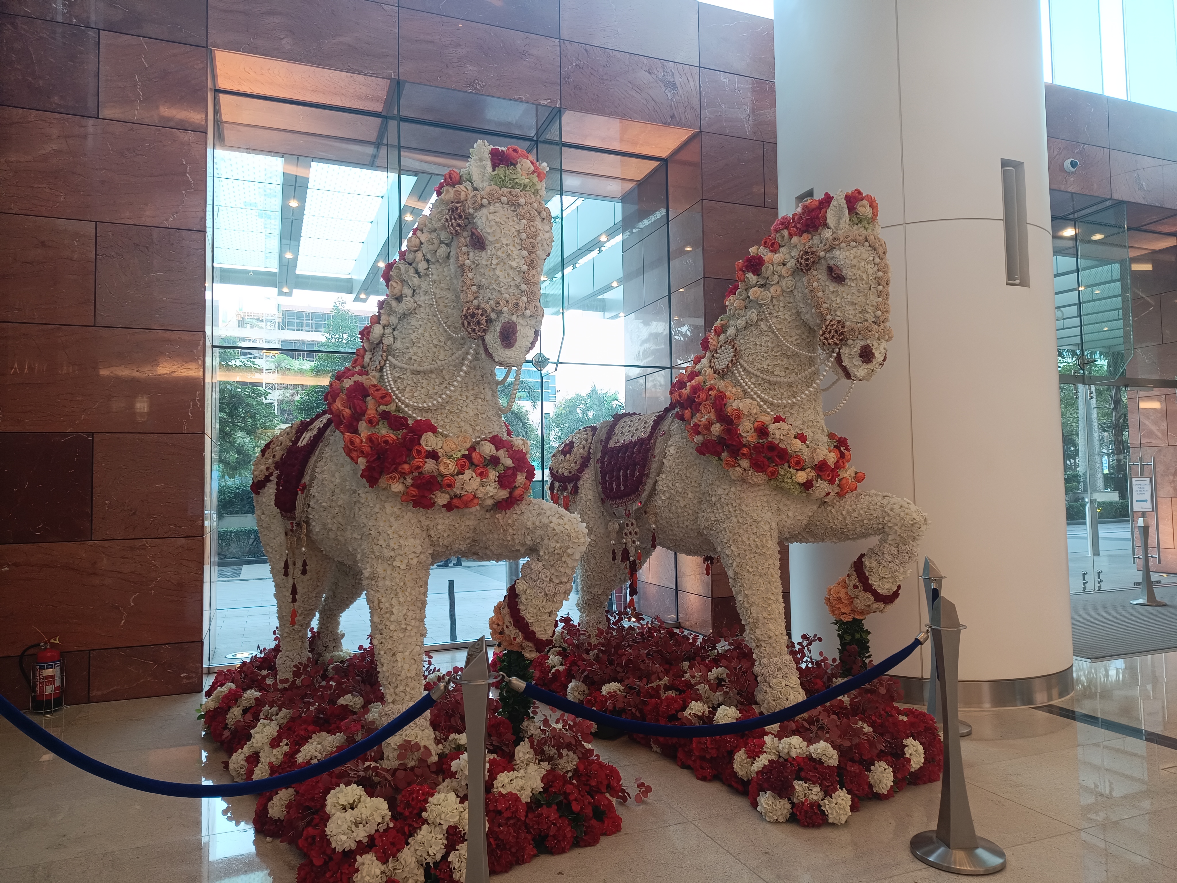 Two majestic horse sculptures made of white and red flowers with floral garlands, displayed in a polished lobby with large glass windows.