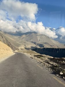 Mountain road under a clear blue sky with fluffy white clouds. The rugged, barren landscape is dotted with hills and distant snow-capped peaks.