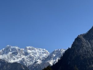 Bright snow covered mountains rise beneath a clear blue sky, while dark forested hills add contrast to the peaceful landscape.