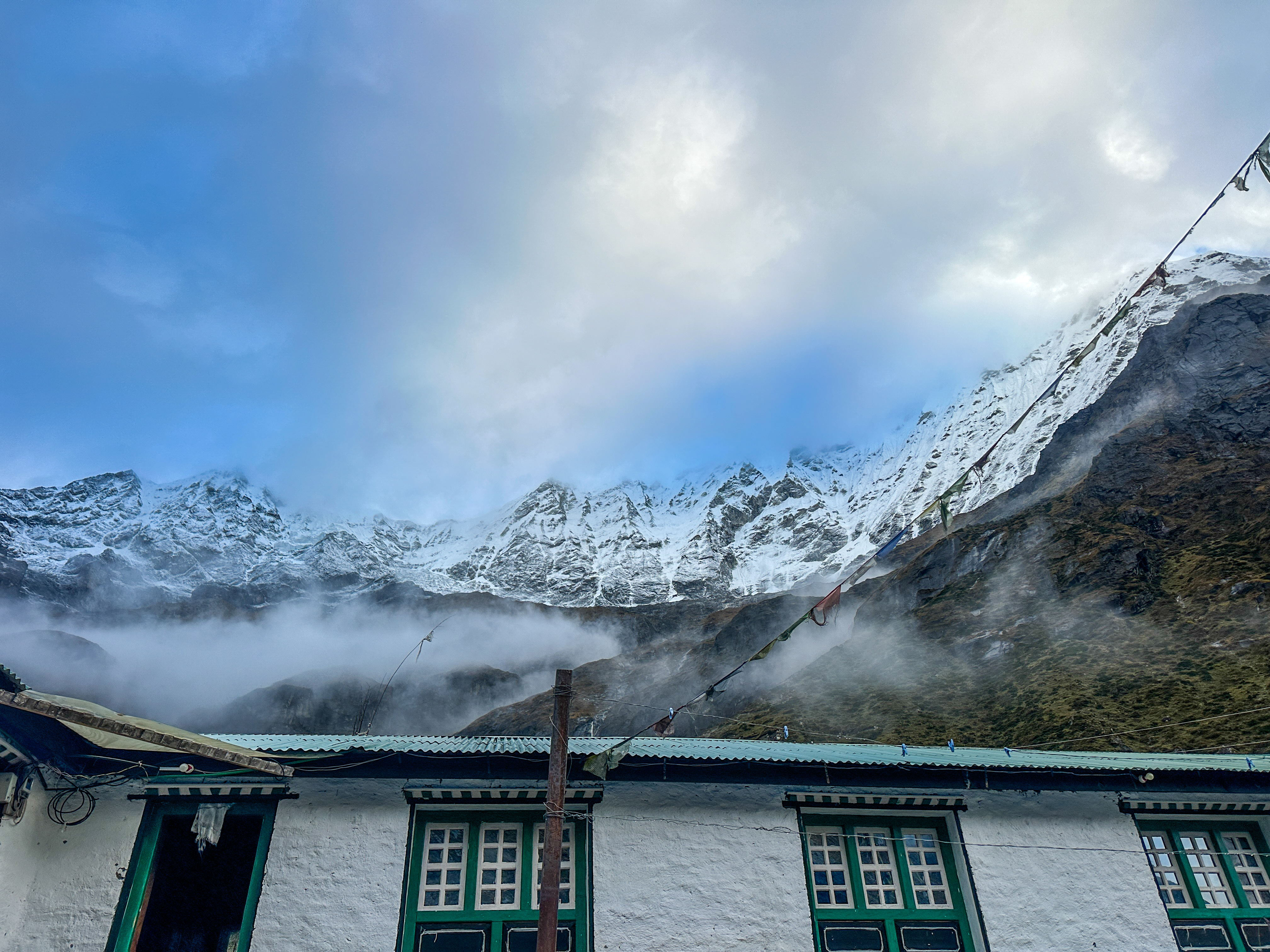 Snow-covered mountains with mist, a cloudy sky, and a building with green windows and prayer flags.
