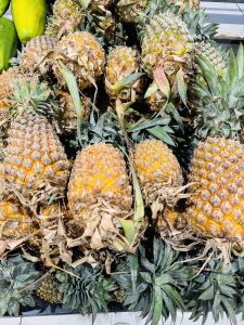 A cluster of ripe, golden-orange pineapples with green spiky leaves and some dried brown leaves, displayed in a market setting.