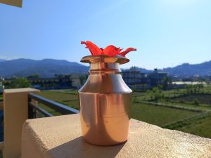 A shiny copper vessel with a red flower placed on a balcony ledge, overlooking green fields, buildings, and distant hills under a clear blue sky.