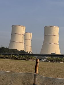 Three large beige cooling towers with red stripes stand in a grassy area near a bridge with light traffic under a soft morning or afternoon sky.