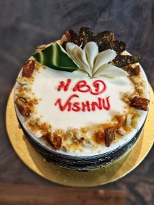 A round cake with white icing, topped with nuts and caramel, has "HBD Vishnu" in red icing.