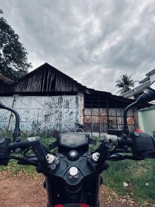 View from a motorcycle facing an old, weathered barn with metal doors under a cloudy sky. 