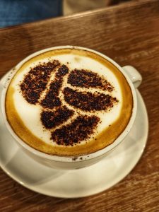 A close-up view of a cup of cappuccino topped with an artistic cocoa powder design resembling leaves, sitting on a white saucer against a wooden surface.