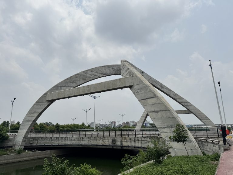 Concrete arch bridge under a cloudy sky, spanning a river with lush greenery.