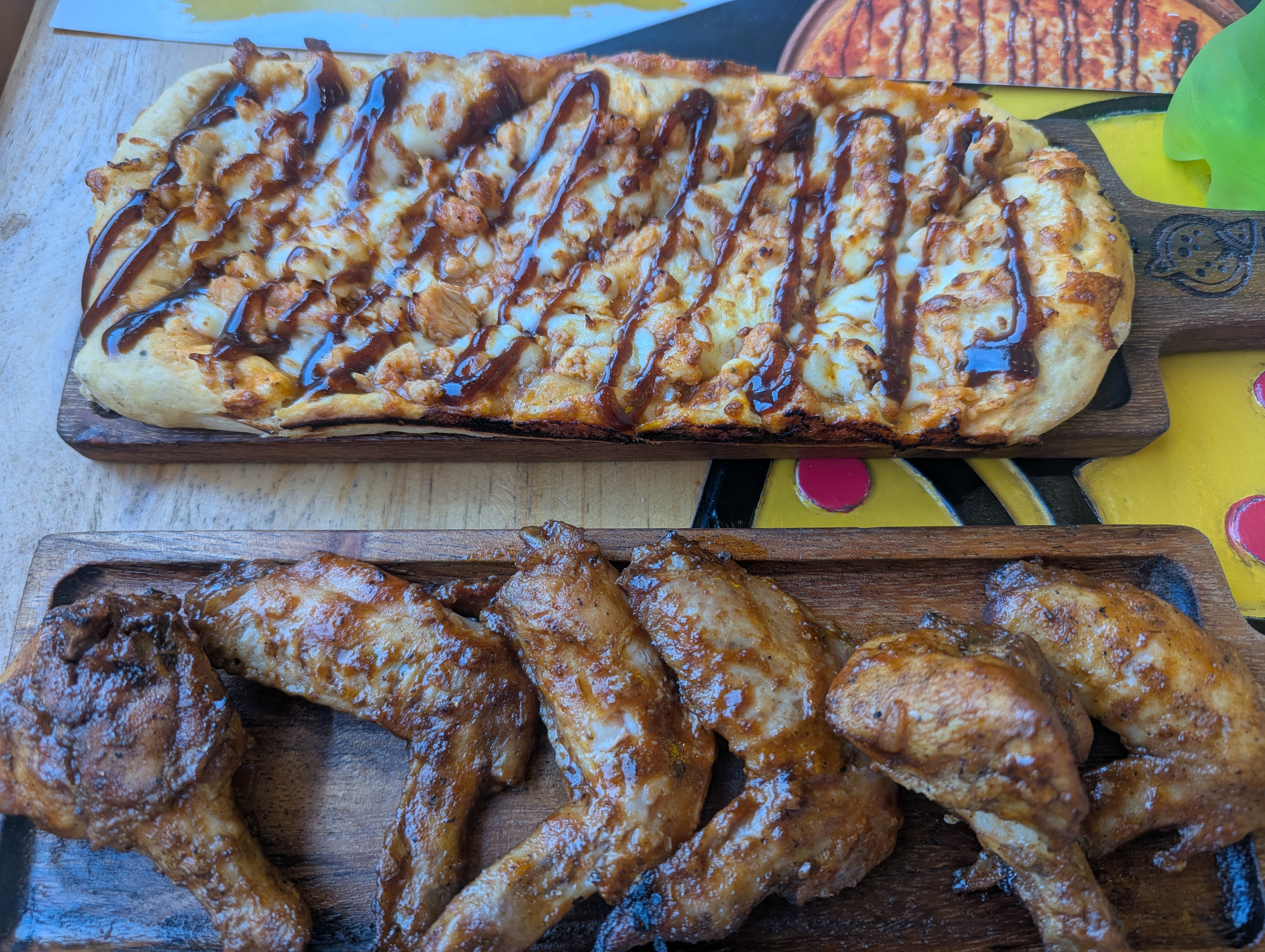 A wooden table has a BBQ chicken pizza with sauce drizzled on top, along with six crispy glazed chicken wings served on a wooden board.